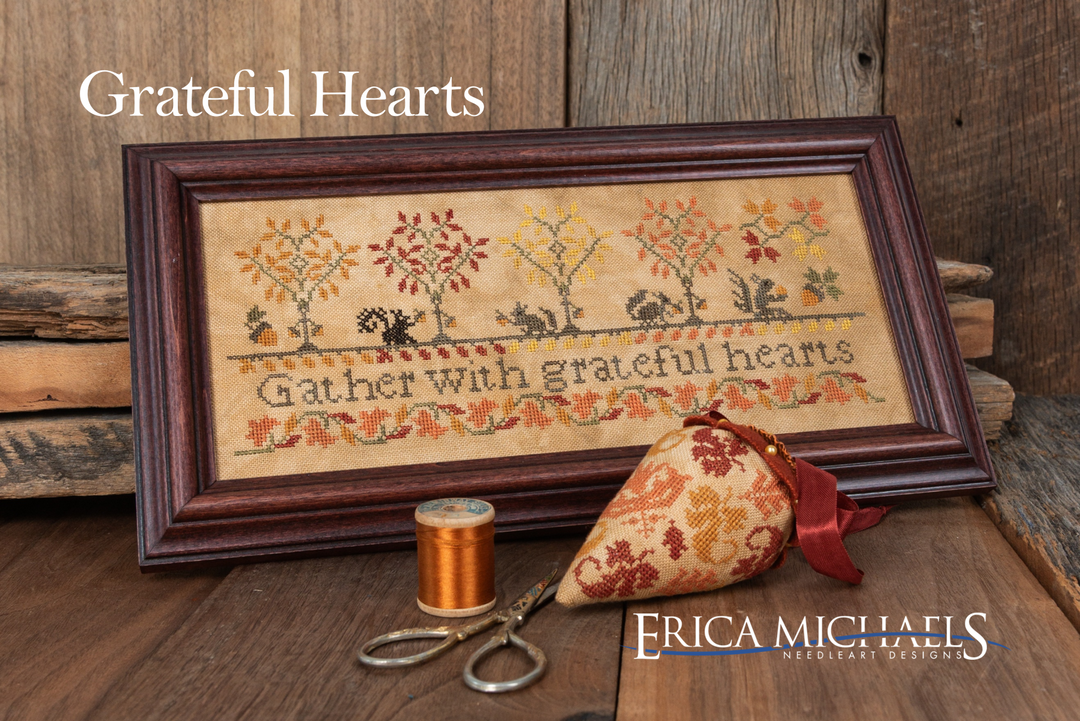 A framed cross-stitch piece reading 'Gather with grateful hearts' with autumn trees and squirrels, displayed on a rustic wooden surface with a spool of thread, scissors, and a fabric ornament. Text at top reads 'Grateful Hearts' and the brand 'Erica Michaels Needleart Designs' appears at the bottom right.
