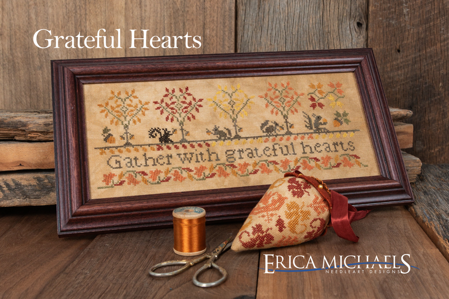 A framed cross-stitch piece reading 'Gather with grateful hearts' with autumn trees and squirrels, displayed on a rustic wooden surface with a spool of thread, scissors, and a fabric ornament. Text at top reads 'Grateful Hearts' and the brand 'Erica Michaels Needleart Designs' appears at the bottom right.