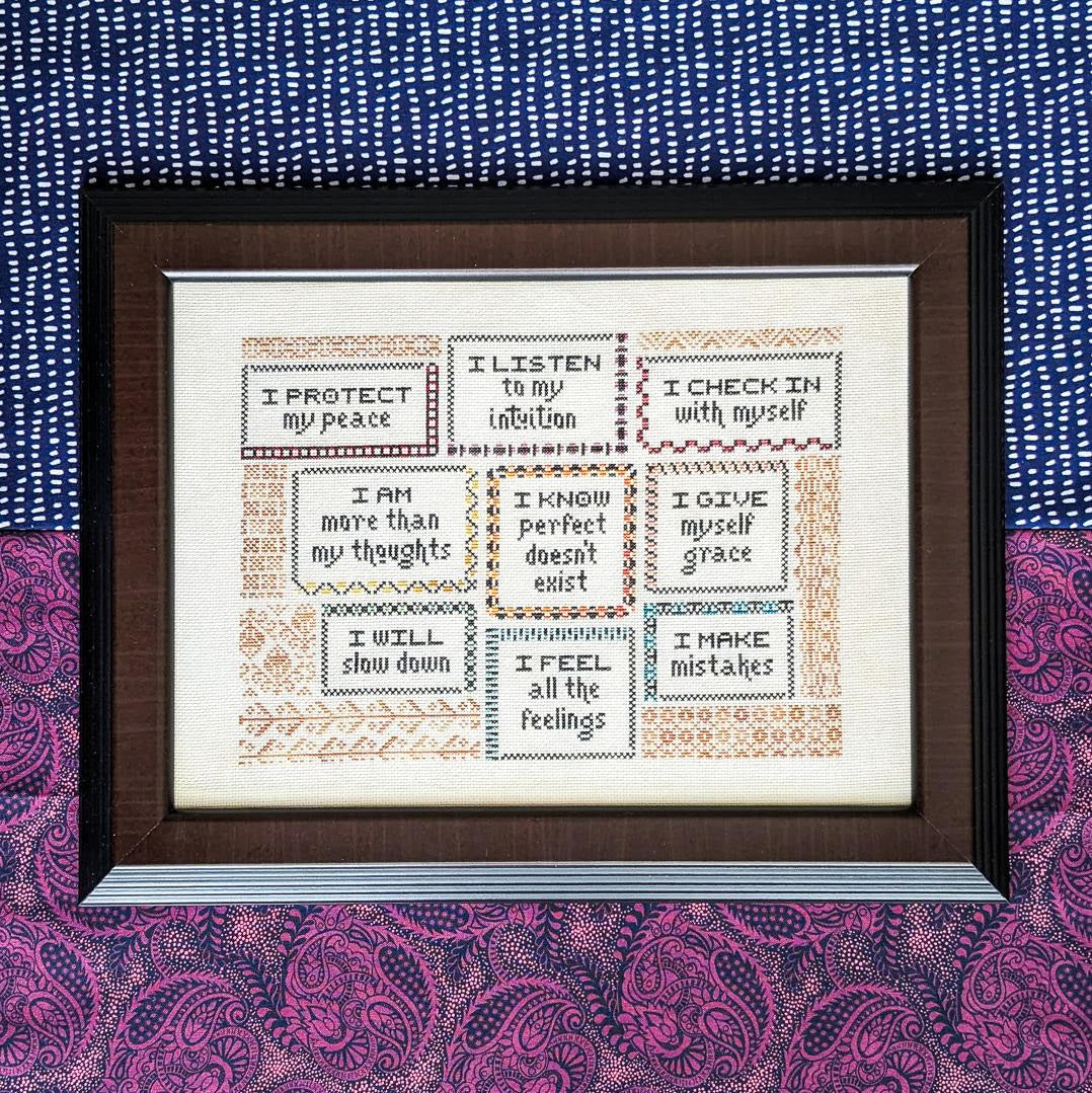 Framed cross-stitched artwork with motivational phrases on a patterned fabric background