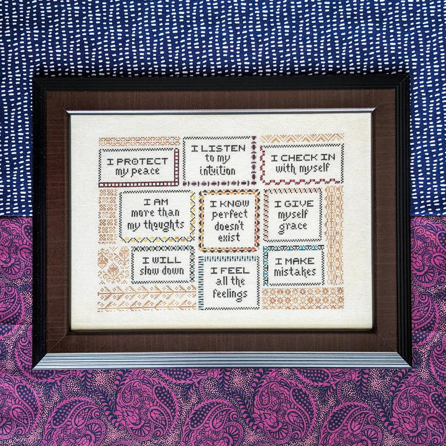 Framed cross-stitched artwork with motivational phrases on a patterned fabric background