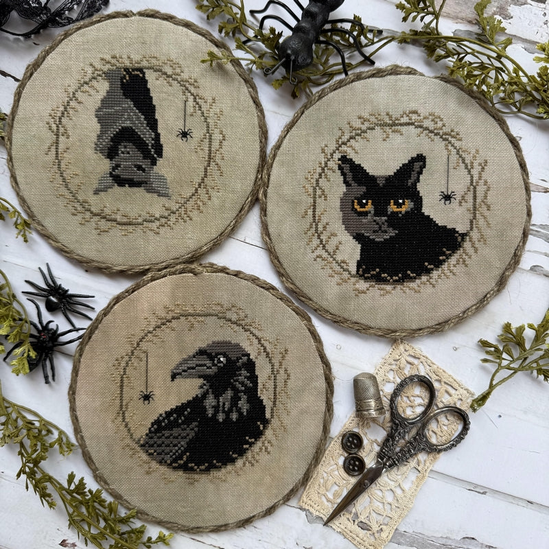 Three round cross-stitch coasters featuring stitched black cat, raven, and bat designs on a beige linen background, arranged on a white wood surface with decorative black spiders, greenery, and scissors.