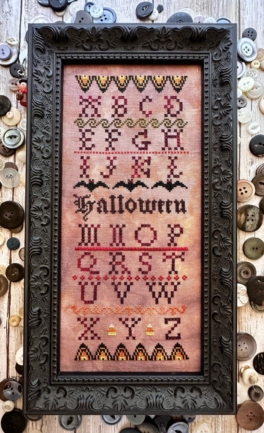 A framed cross-stitch sampler featuring the word 'Halloween' in stylized lettering, surrounded by decorative buttons on a rustic wood background.