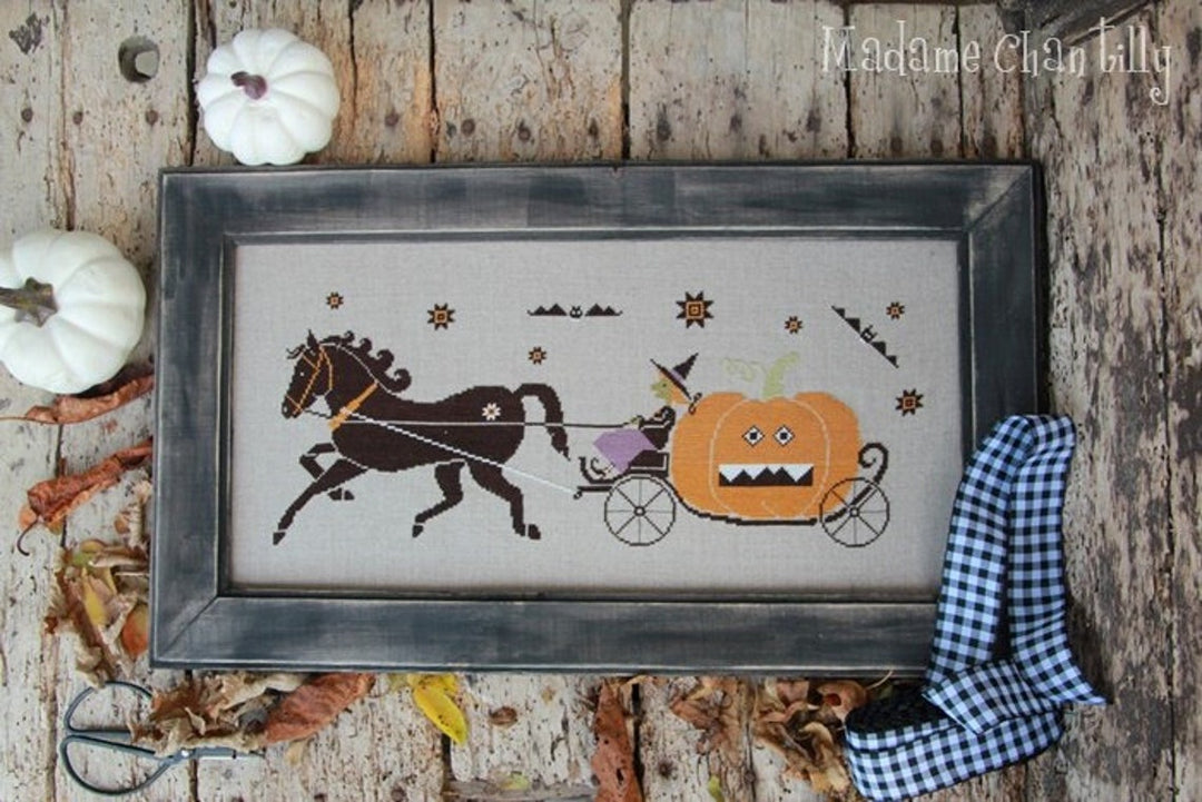 Framed Halloween-themed cross-stitch with a horse, pumpkin carriage, and decorative ribbon on a wooden background.