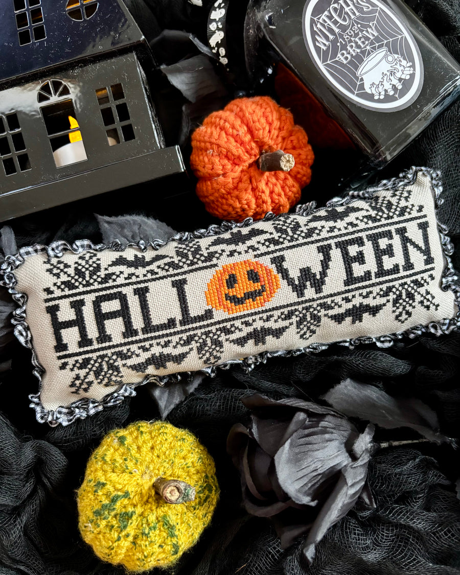 A Halloween-themed cross-stitch pillow with 'HALLOWEEN' and a jack-o'-lantern motif, styled with miniature pumpkins, black lace, a house lantern, and a bottle of Witch's Brew on a dark fabric background.