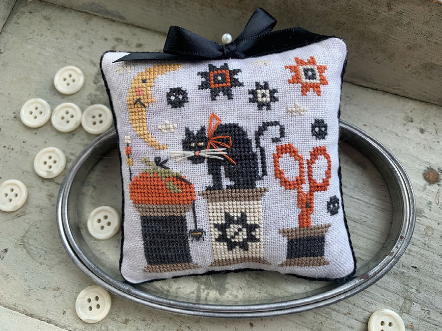 A square cross-stitch pillow with a black ribbon bow on top, featuring a Halloween-themed design with a black cat, orange pumpkin, scissors, and stars on a white background. The pillow is styled on a silver metal tray with decorative buttons on a rustic stone surface.