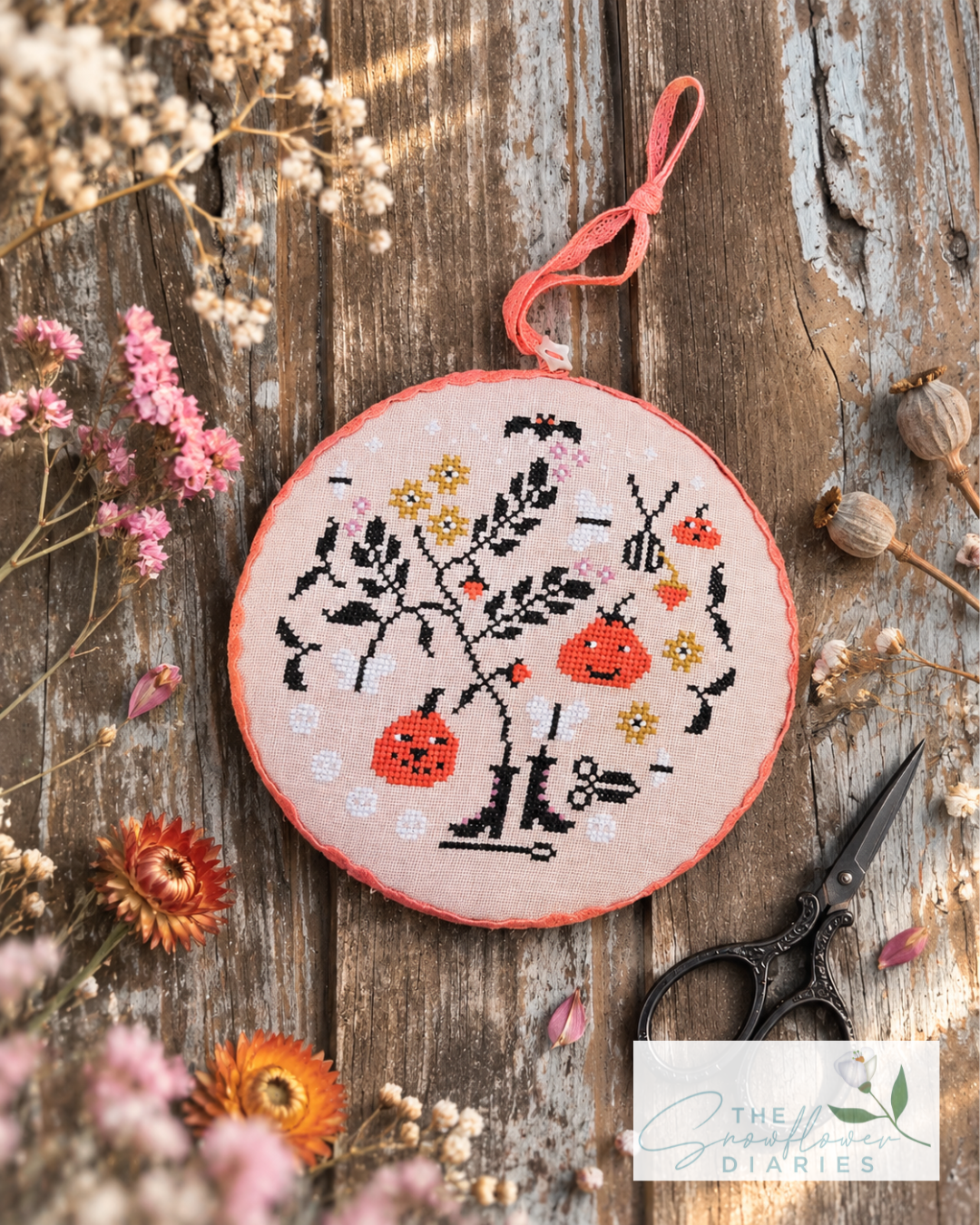 A circular embroidered cross-stitch ornament in a wooden hoop, featuring a whimsical design of red pumpkins, black boots, and yellow flowers on a pink fabric background. The hoop is displayed on a rustic wooden surface surrounded by dried flowers, with a pair of embroidery scissors and a logo for 'The Needlework Diaries' in the corner.
