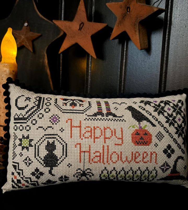 A rectangular cross-stitch pillow with a black pom-pom border, featuring a 'Happy Halloween' design with a black cat, witch hat, pumpkin, and crow on a cream fabric background. The pillow is styled on a dark paneled wall next to a lit candle and rustic wooden star decorations.