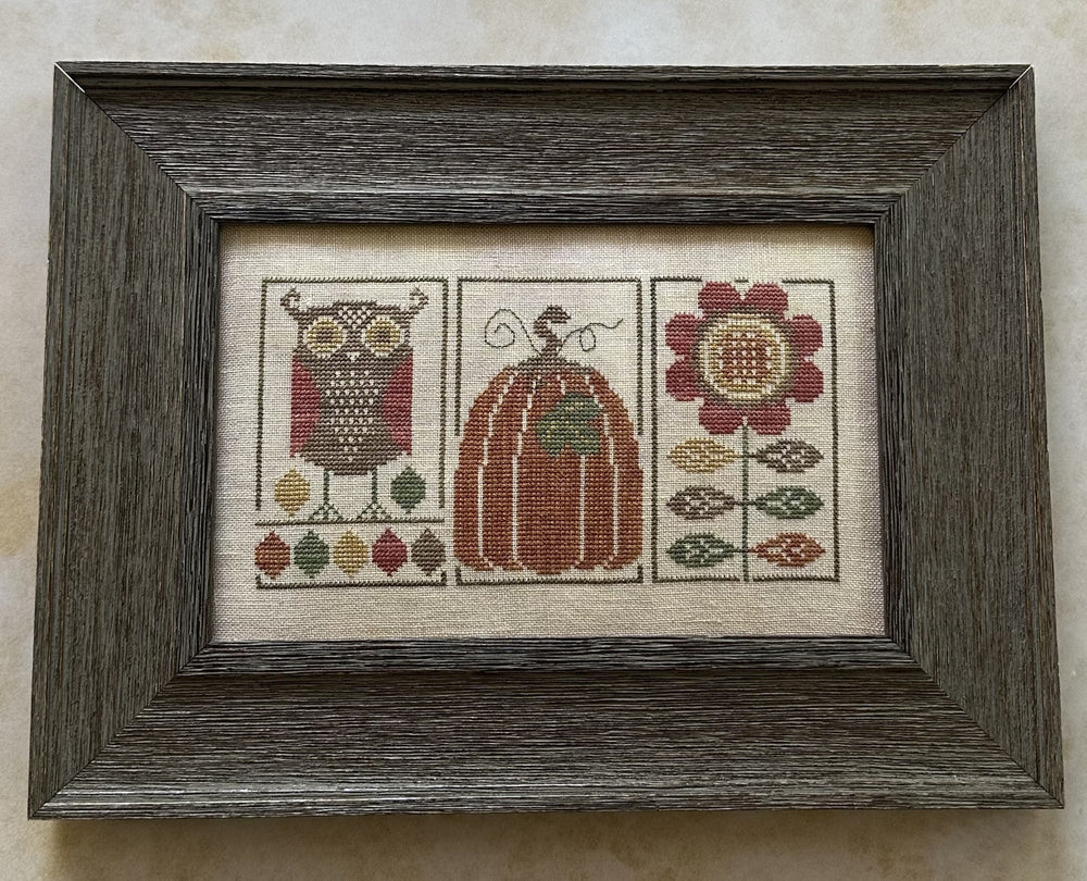 Framed cross-stitched piece with owl, pumpkin, and flower design on a beige background
