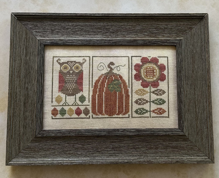 Framed cross-stitched piece with owl, pumpkin, and flower design on a beige background