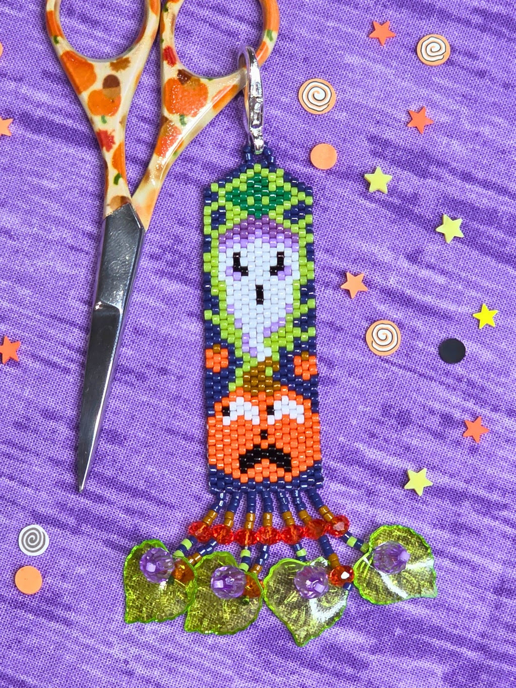 A handmade beaded keychain featuring a ghost and pumpkin design, displayed on a purple textured background with decorative paper stars and orange-handled scissors.