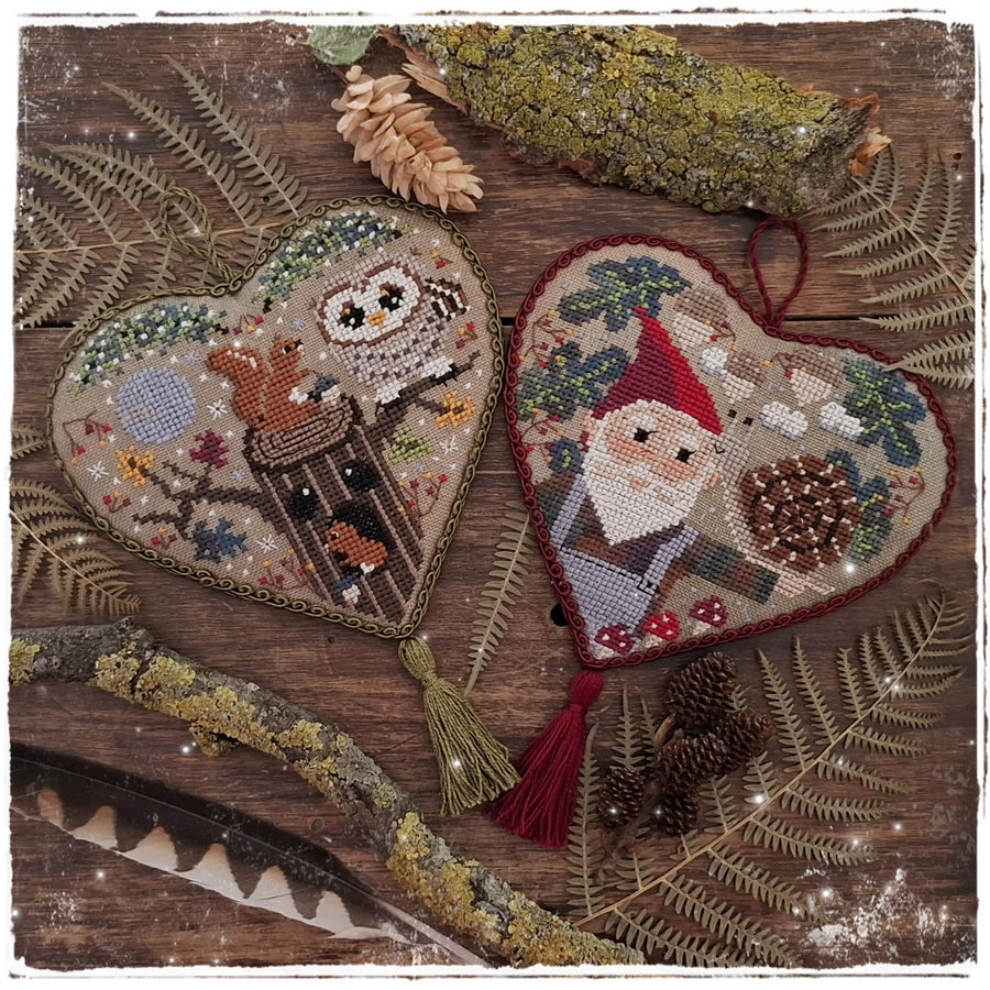 Two heart-shaped cross-stitch ornaments displayed on a rustic wooden surface with pinecones, ferns, and mossy branches. One ornament features an owl perched on a tree trunk, and the other shows a Santa Claus face with a red hat and beard.