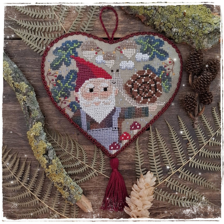 Heart-shaped embroidered cross-stitch ornament featuring a gnome, deer, pinecone, and mushrooms, displayed on a rustic wooden background with ferns and moss.