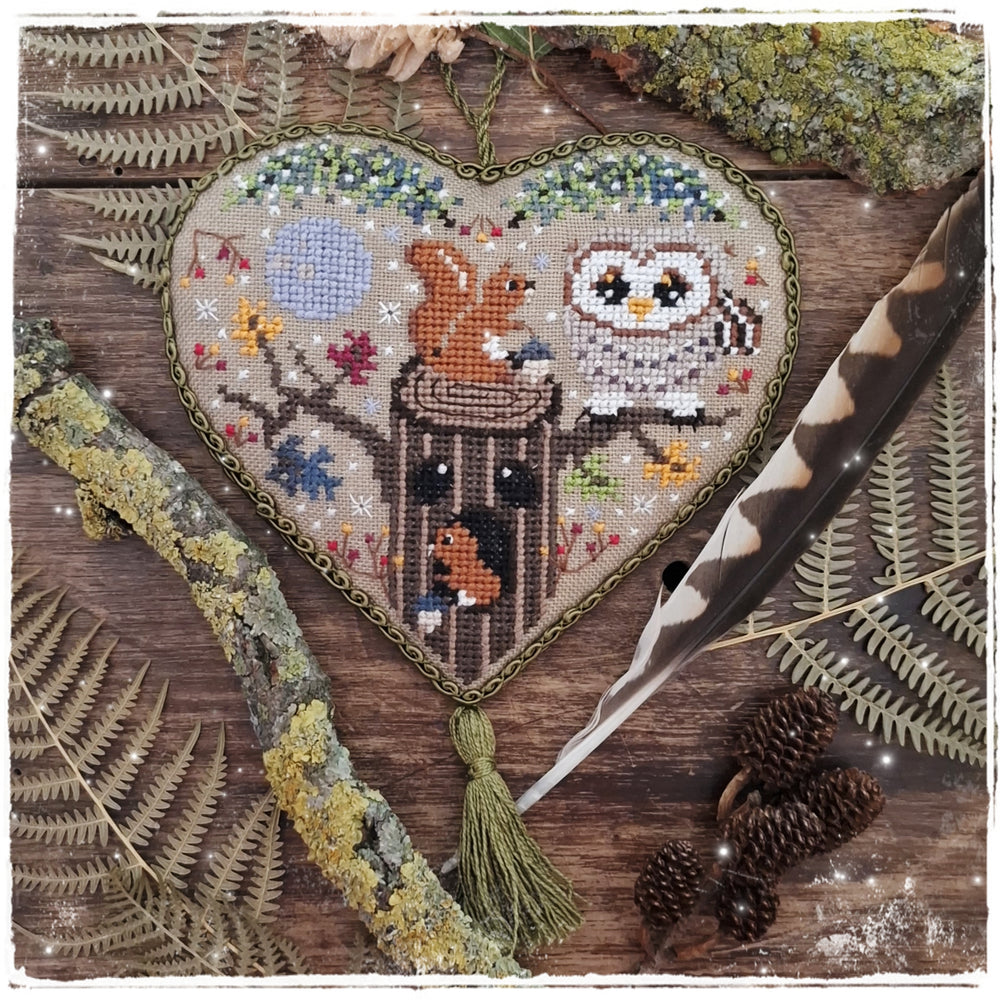 Heart-shaped cross-stitch hanging ornament featuring a woodland scene with an owl, squirrel, birdhouse, and autumn leaves, displayed on a rustic wooden surface with ferns, moss, a feather quill, and pinecones.
