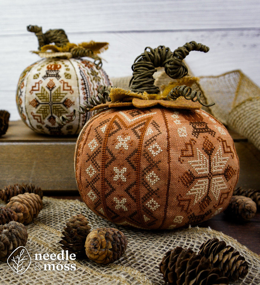Two decorative cross-stitch pumpkins displayed on a rustic surface with burlap, pinecones, and a wooden box in the background. The foreground pumpkin is rust orange with cream and brown geometric and snowflake patterns, and the background pumpkin is cream with multicolor patterns.