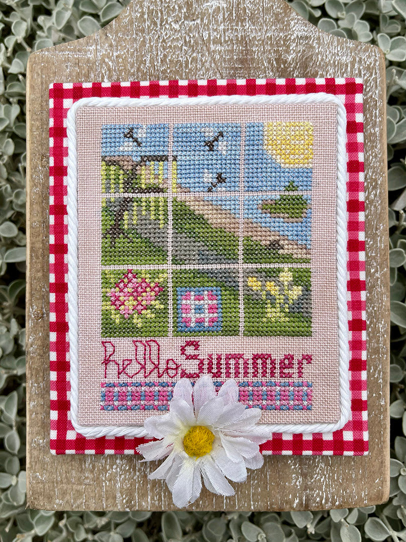 A decorative cross-stitch piece mounted on a rustic wooden plaque, featuring a 3x3 summer scene with a windmill, house, flowers, and the words 'Hello Summer' in pink embroidery, accented with a white daisy flower in front.