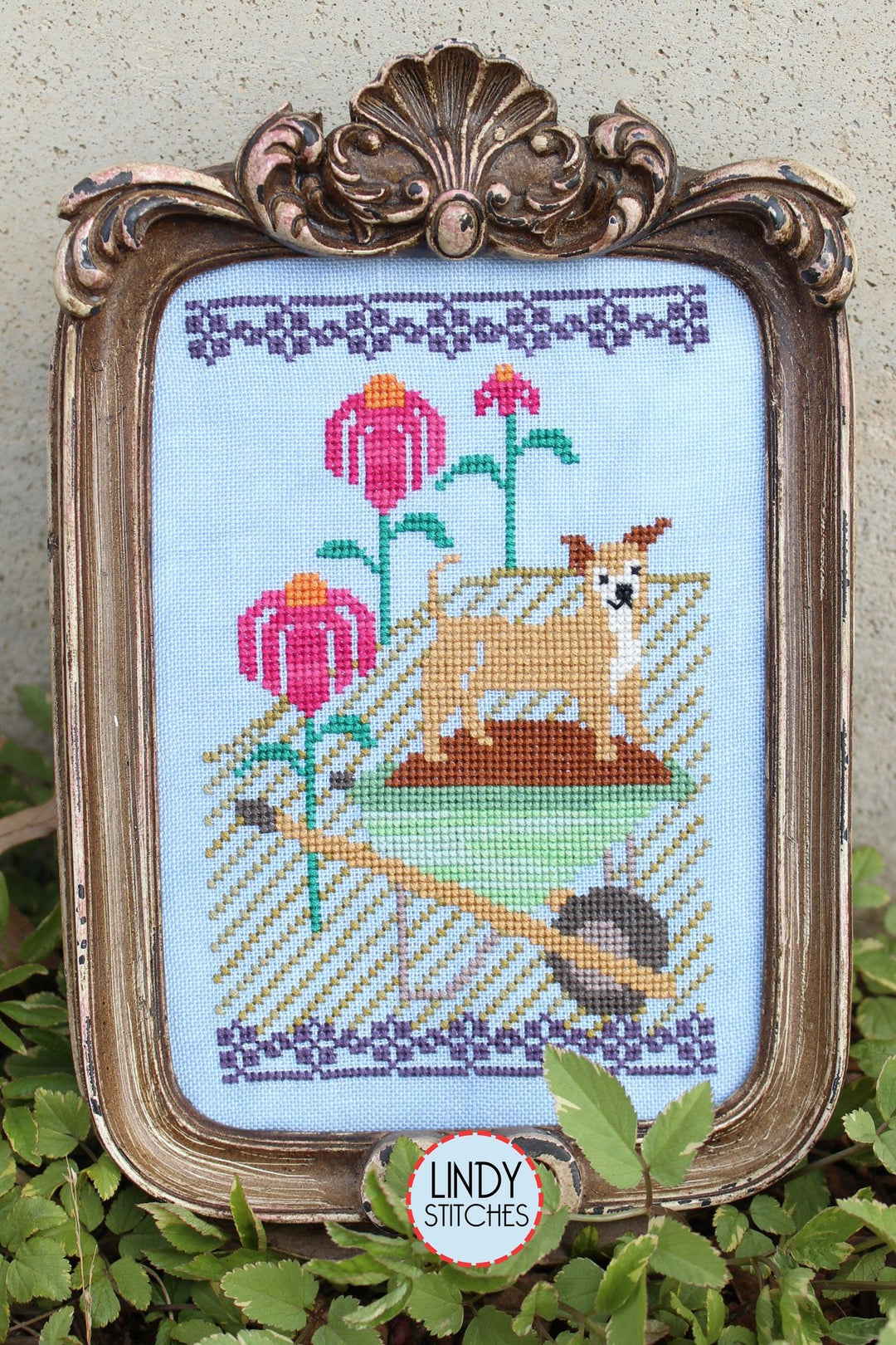 Framed cross-stitched piece with a dog and flowers, featuring Lindy Stitches branding.