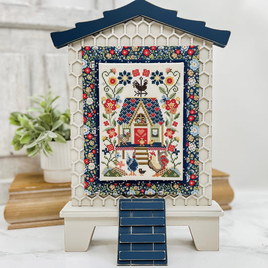 Decorative quilt with floral patterns and a house design on a stand with a white background.