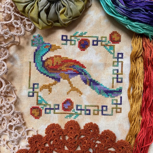 Cross-stitched peacock design on fabric with colorful threads and lace.