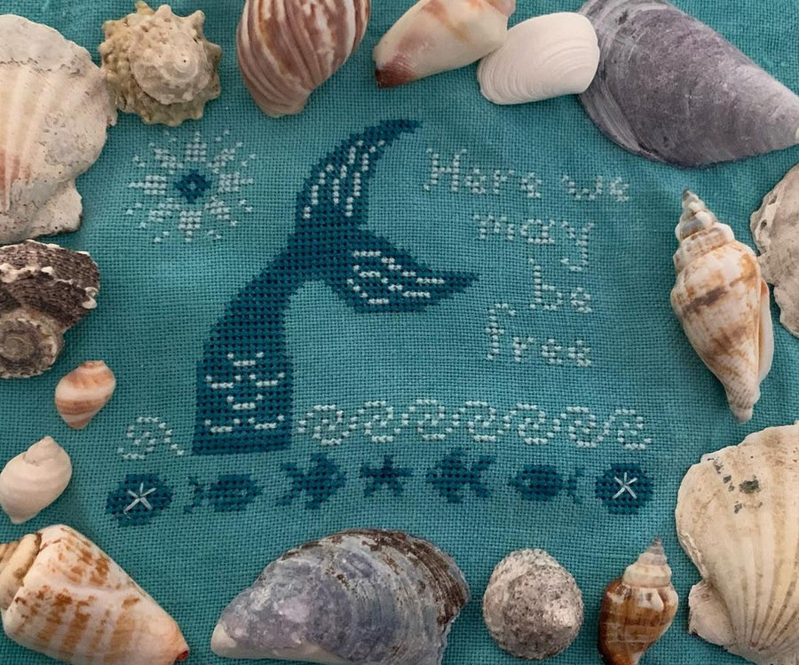 Cross-stitched mermaid design on teal fabric with seashells around it.