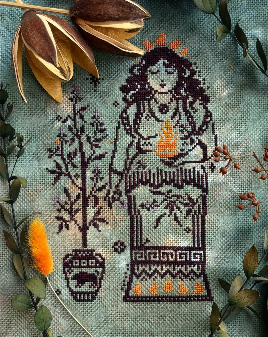 A cross-stitch or needlepoint textile artwork on a muted blue-green fabric background, featuring a stylized woman holding a flame, a potted tree, and geometric border details. The piece is styled with dried botanicals and greenery around the edges.