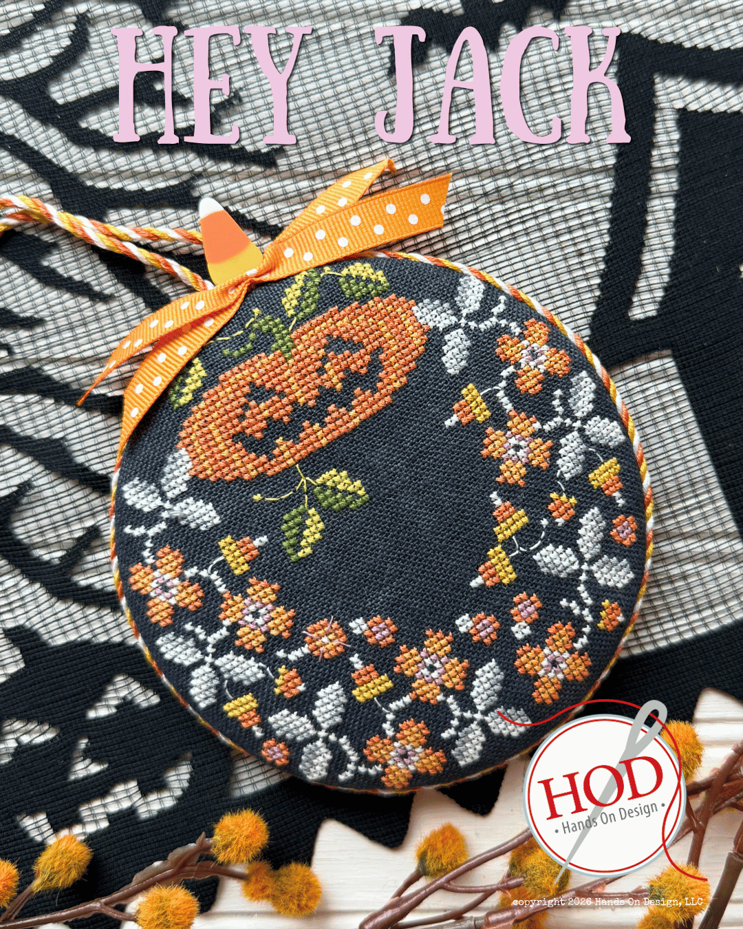 A round cross-stitch sampler featuring a Halloween design with a pumpkin, floral wreath, and spiderweb pattern on a black fabric background, tied with an orange polka-dot ribbon. The text 'HEY JACK' is written at the top, and a 'HOD' logo is visible in the bottom right corner.