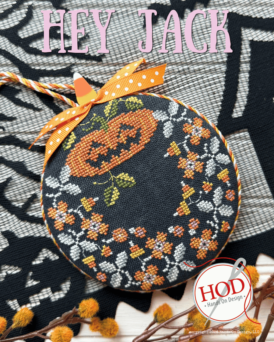 A round cross-stitch sampler featuring a Halloween design with a pumpkin, floral wreath, and spiderweb pattern on a black fabric background, tied with an orange polka-dot ribbon. The text 'HEY JACK' is written at the top, and a 'HOD' logo is visible in the bottom right corner.