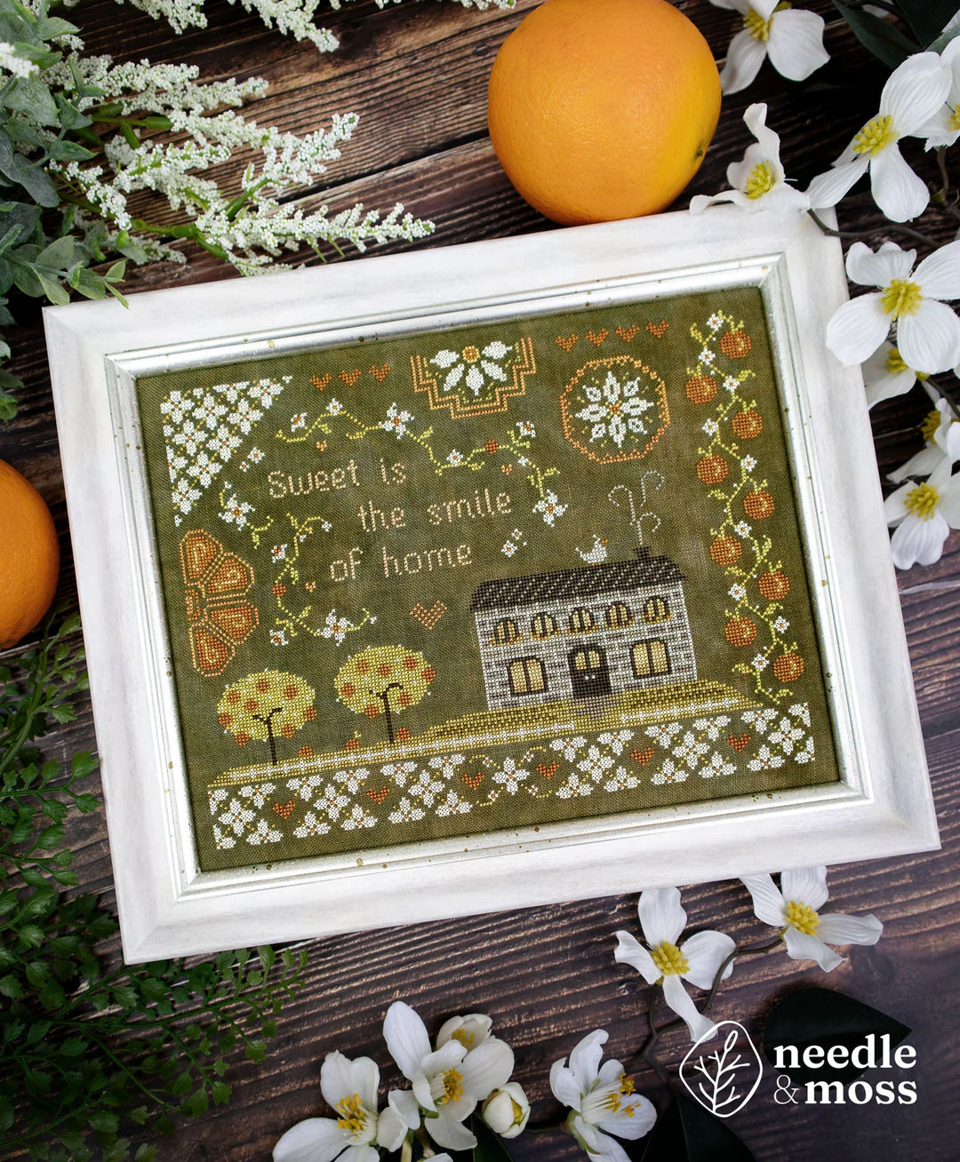 A framed needlework sampler with a rustic country house design and the text 'Sweet is the smile of home,' styled on a dark wood surface with white flowers, greenery, and oranges. The Needle & Moss logo appears in the lower right corner.
