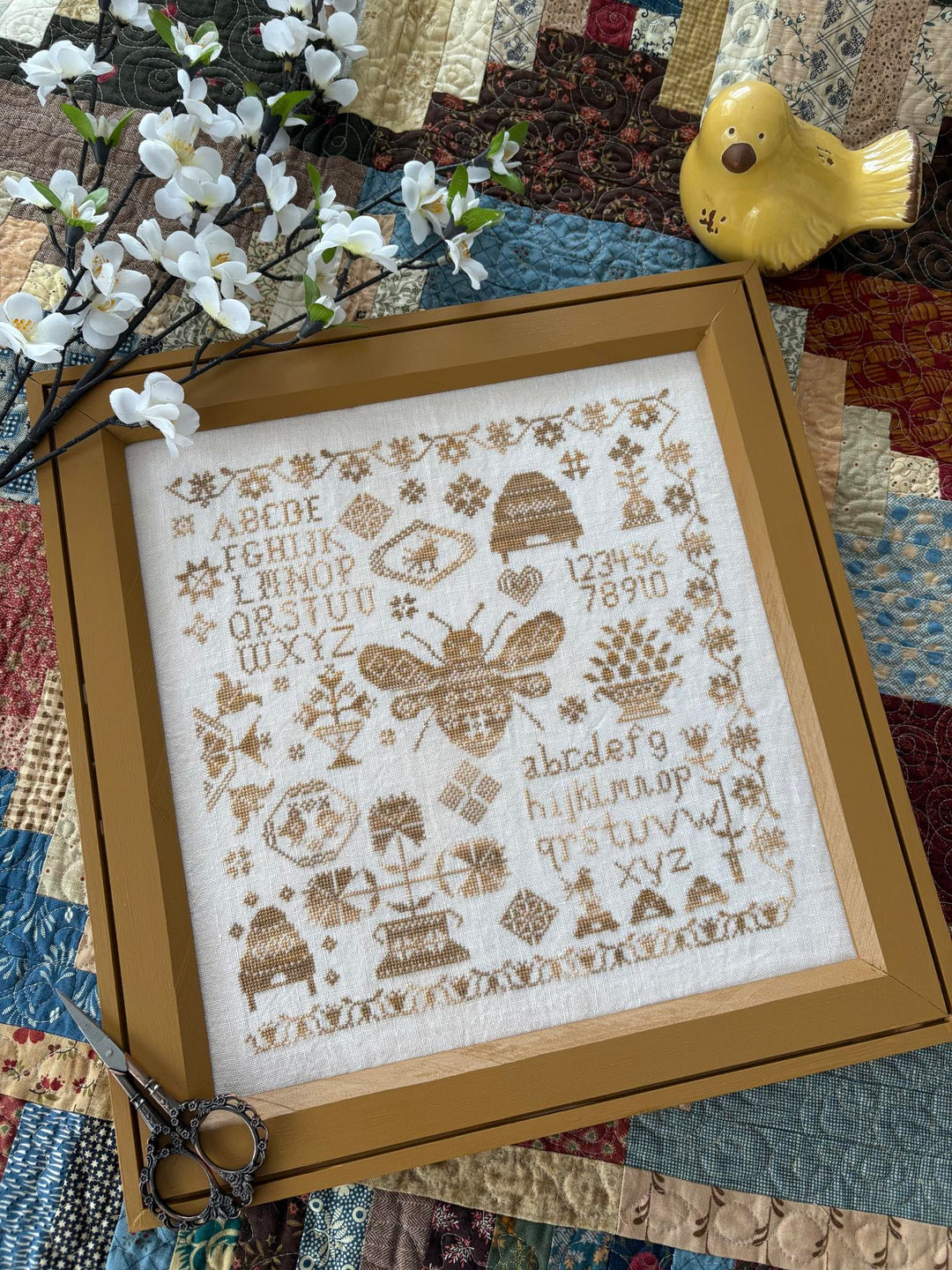 Framed cross-stitched sampler with bees and alphabets on a quilted background.