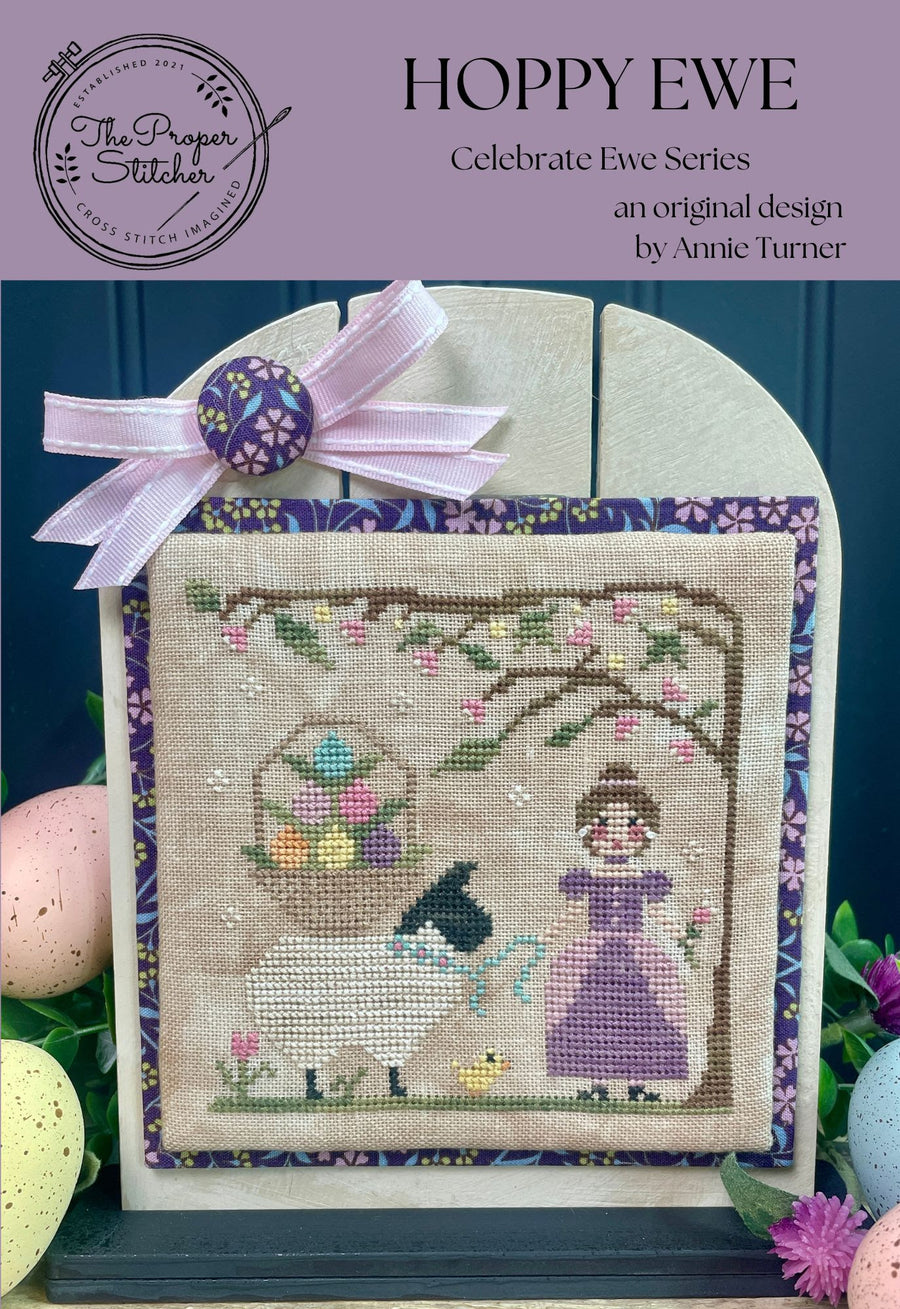 Cross-stitched design of a woman with a lamb and Easter eggs, framed with a pink bow, on a dark blue background.