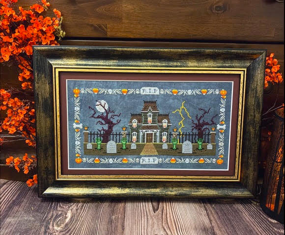 A framed Halloween-themed needlepoint or cross-stitch artwork featuring a haunted house, tombstones, lightning, and autumn decorations, displayed on a wooden surface with orange foliage in the background.