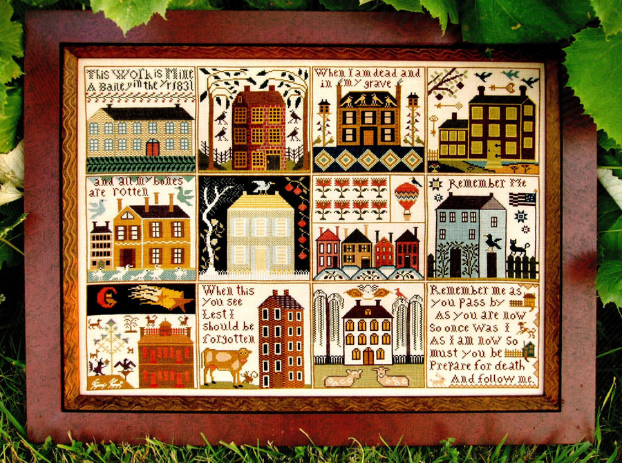 Framed cross-stitch sampler with various buildings and text on a grass background