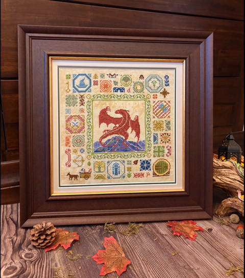 A framed cross-stitch artwork featuring a central red dragon over blue waves, surrounded by colorful decorative motifs, displayed in a dark wood frame on a rustic wooden surface with autumn leaves and logs nearby.