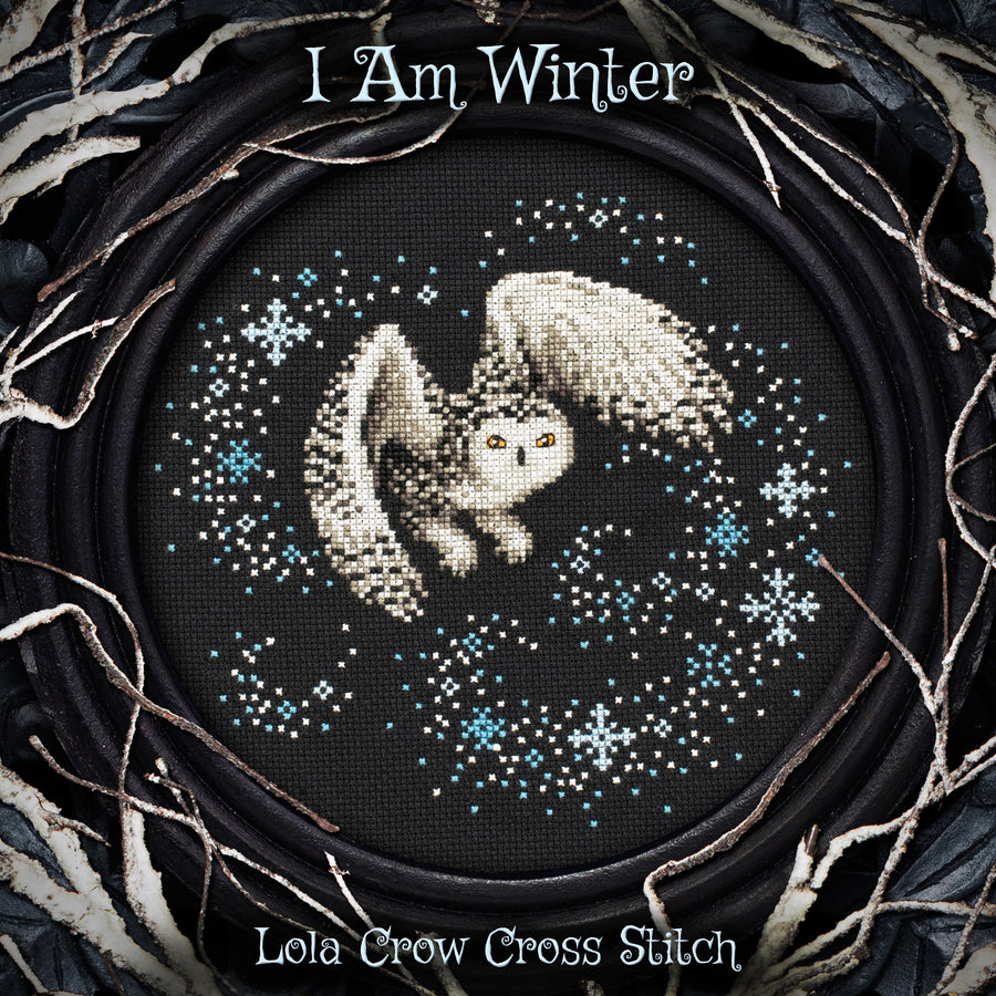 A circular cross stitch artwork featuring a white owl with wings spread, surrounded by blue and white snowflakes on a black background. The stitched design is framed by a dark circular border with branches, and the text 'I Am Winter' appears at the top and 'Lola Crow Cross Stitch' at the bottom.