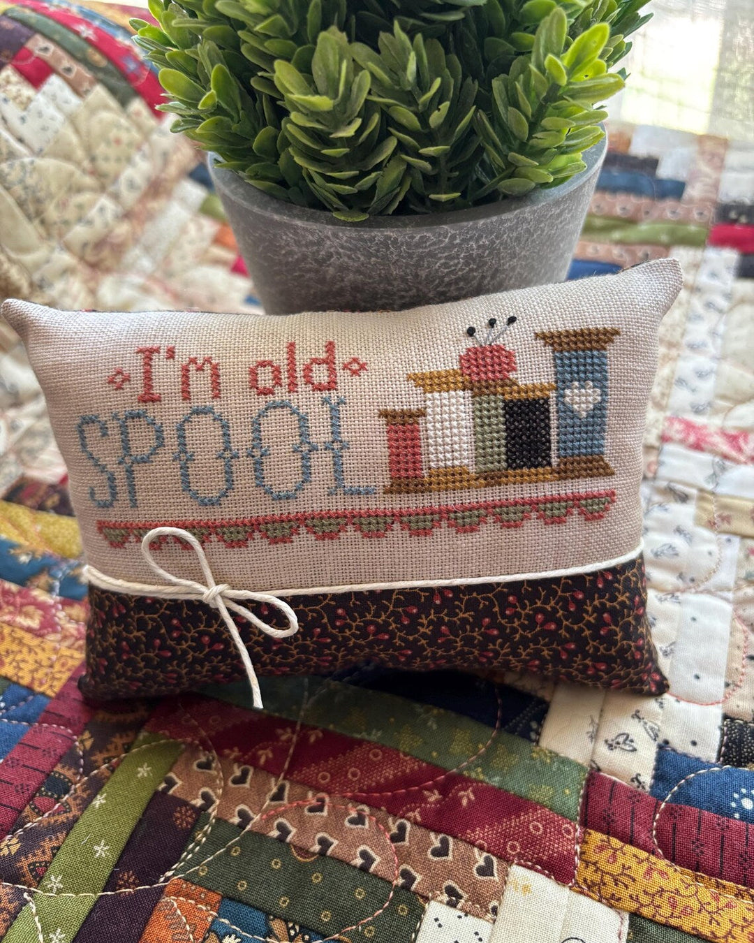 A small embroidered cross-stitch pillow with a floral fabric base and a beige top panel reading “I’m old SPOON” with a small illustrated spool motif, styled on a quilted blanket with a potted green plant behind it.
