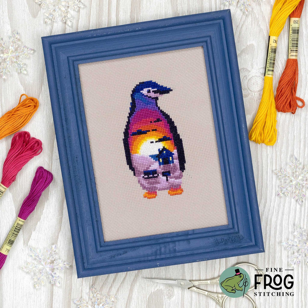 A framed cross-stitch design of a penguin with a colorful sunset pattern inside its body, set in a blue frame on a white wood background with embroidery floss and scissors.