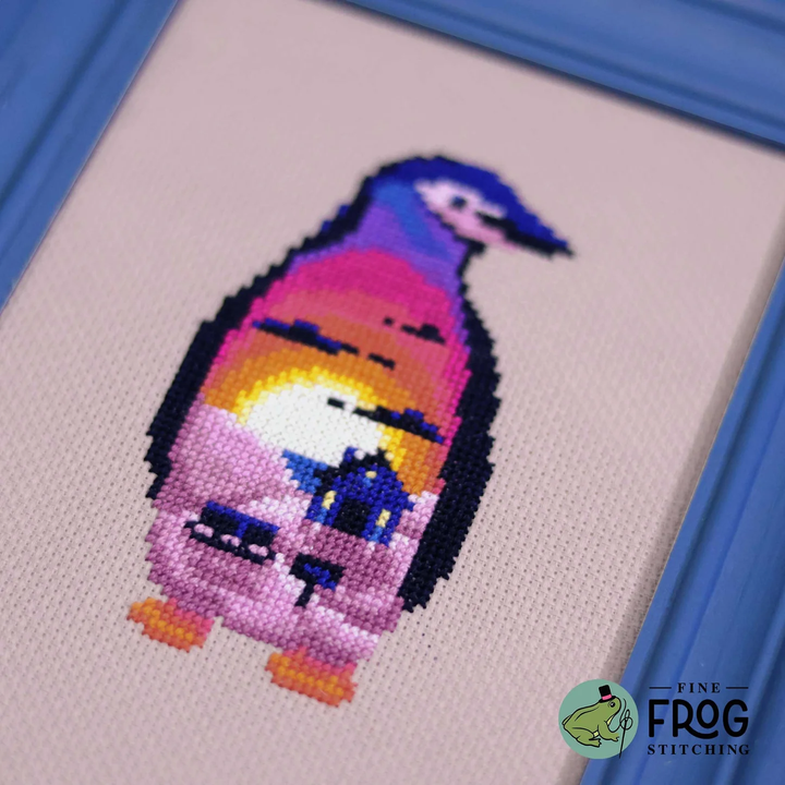 A close-up of a framed cross-stitch artwork showing a stylized penguin with a colorful sunset gradient on its body, bordered by a blue frame. The watermark in the corner reads Fine Frog Stitching.