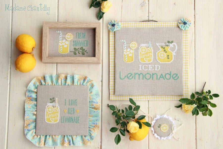 A collection of cross stitch patterns with lemonade designs, including the text 'iced lemongrass' and images of lemonades, displayed on a wooden surface with lemons and flowers around.