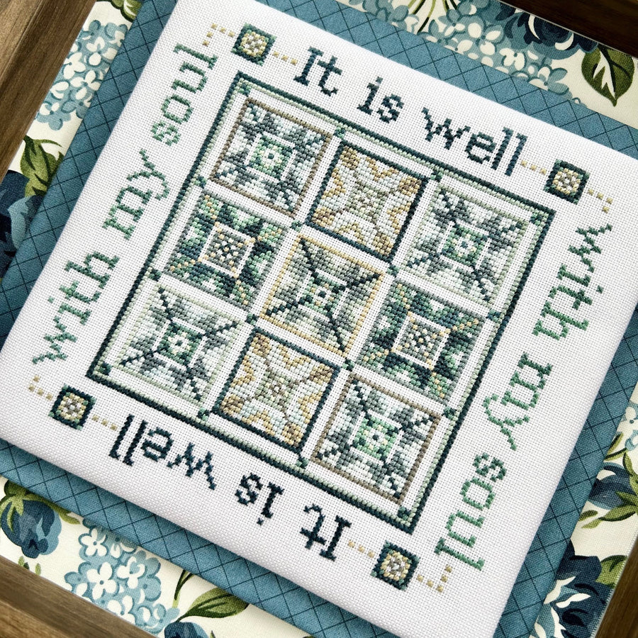 Cross-stitched sampler with text 'It is well with my soul' on a floral fabric background