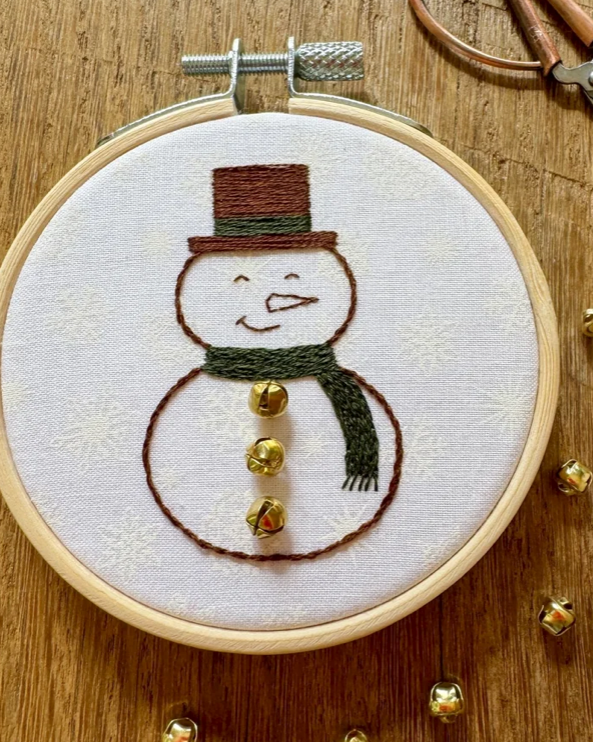 A close-up of a finished embroidery project in a wooden hoop, featuring a snowman with a brown top hat, green scarf, and gold buttons on a white fabric background, set on a wooden surface with scattered gold jingles.