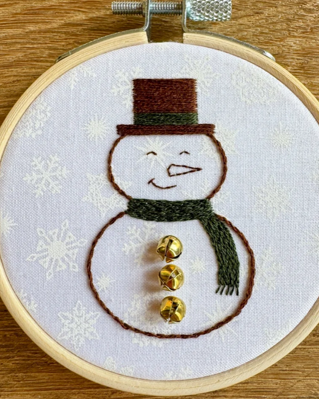 A circular embroidery hoop holding white fabric with a stitched snowman design. The snowman has a brown top hat, dark green scarf, and three shiny gold bells on its body.