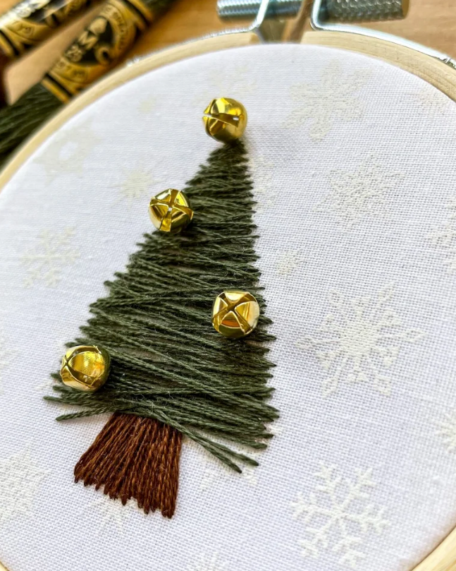 A close-up of a hand-embroidered Christmas tree in olive green thread with four shiny gold bells and a brown trunk, stitched inside a wooden embroidery hoop on white fabric with embroidered snowflakes.