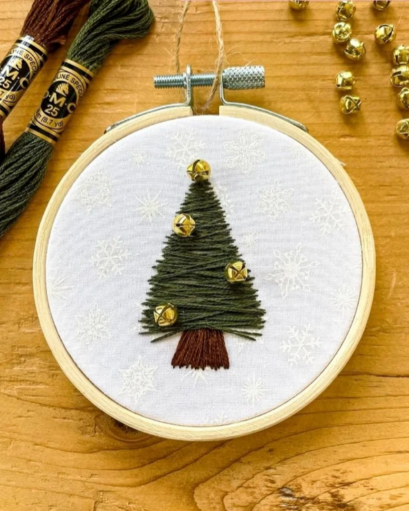 A round embroidery hoop displays a completed Christmas tree design made from dark green thread with small gold bells and a brown trunk, set on a wooden surface with embroidery floss and gold bells nearby.