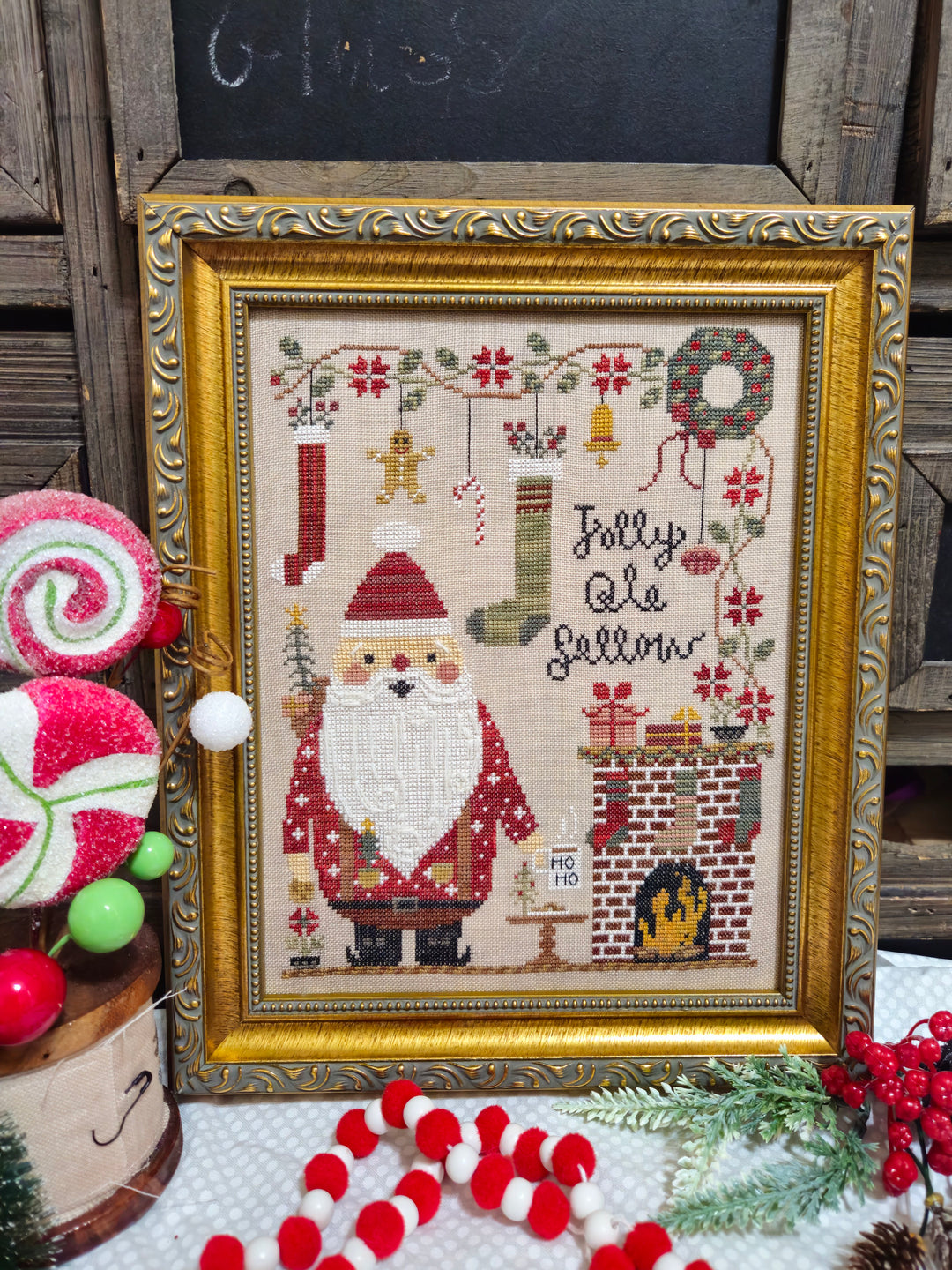 Framed cross-stitched picture of Santa Claus with decorative Christmas elements on a rustic background.