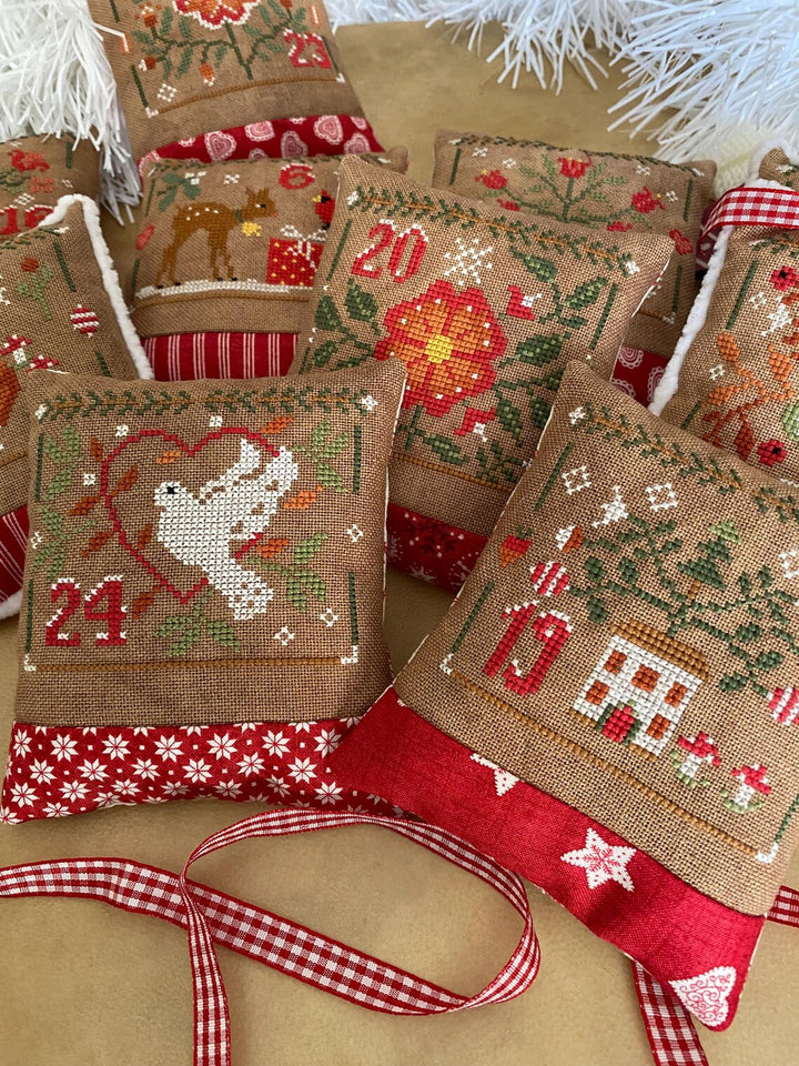 A group of handmade burlap Christmas pillow ornaments with red gingham ribbon trim, featuring embroidered holiday motifs including a dove, a house, a tree, and numbers on a tan background.