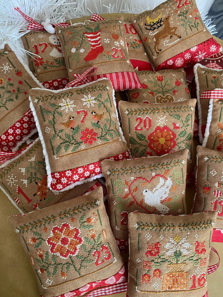 A pile of handmade Christmas cross-stitch cushion ornaments made from tan burlap fabric, decorated with colorful floral and holiday motifs, red and white striped trim, and small hanging ribbons.