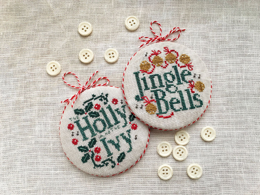 Two round cross-stitch Christmas ornaments on a textured beige linen background, surrounded by decorative buttons. One ornament reads 'Holly, Ivy, 1914' in green and red, and the other reads 'Jingle Bells' in green and red with gold accents.