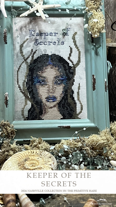 Framed cross-stitched art of a woman with horns titled 'Keeper of the Secrets' on a decorative background.
