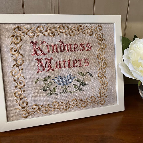 Framed cross-stitched artwork with 'Kindness Matters' text on a wooden surface.