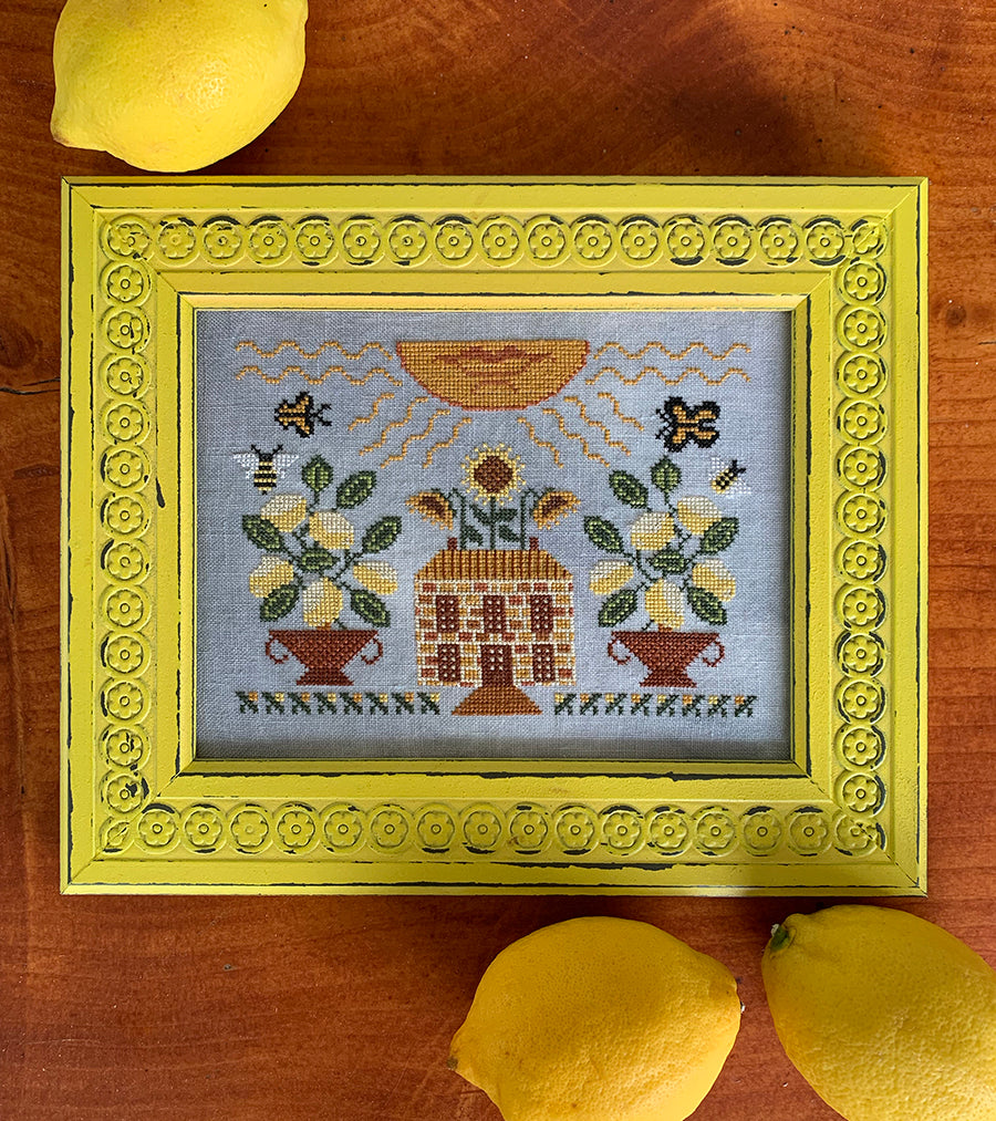 Decorative framed needlepoint with a house, flowers, and bees on a wooden surface with lemons.