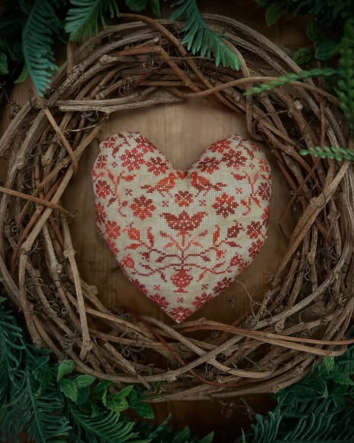 A heart-shaped decorative pillow with a red floral pattern on a cream fabric background, nestled inside a rustic twig wreath with green foliage.